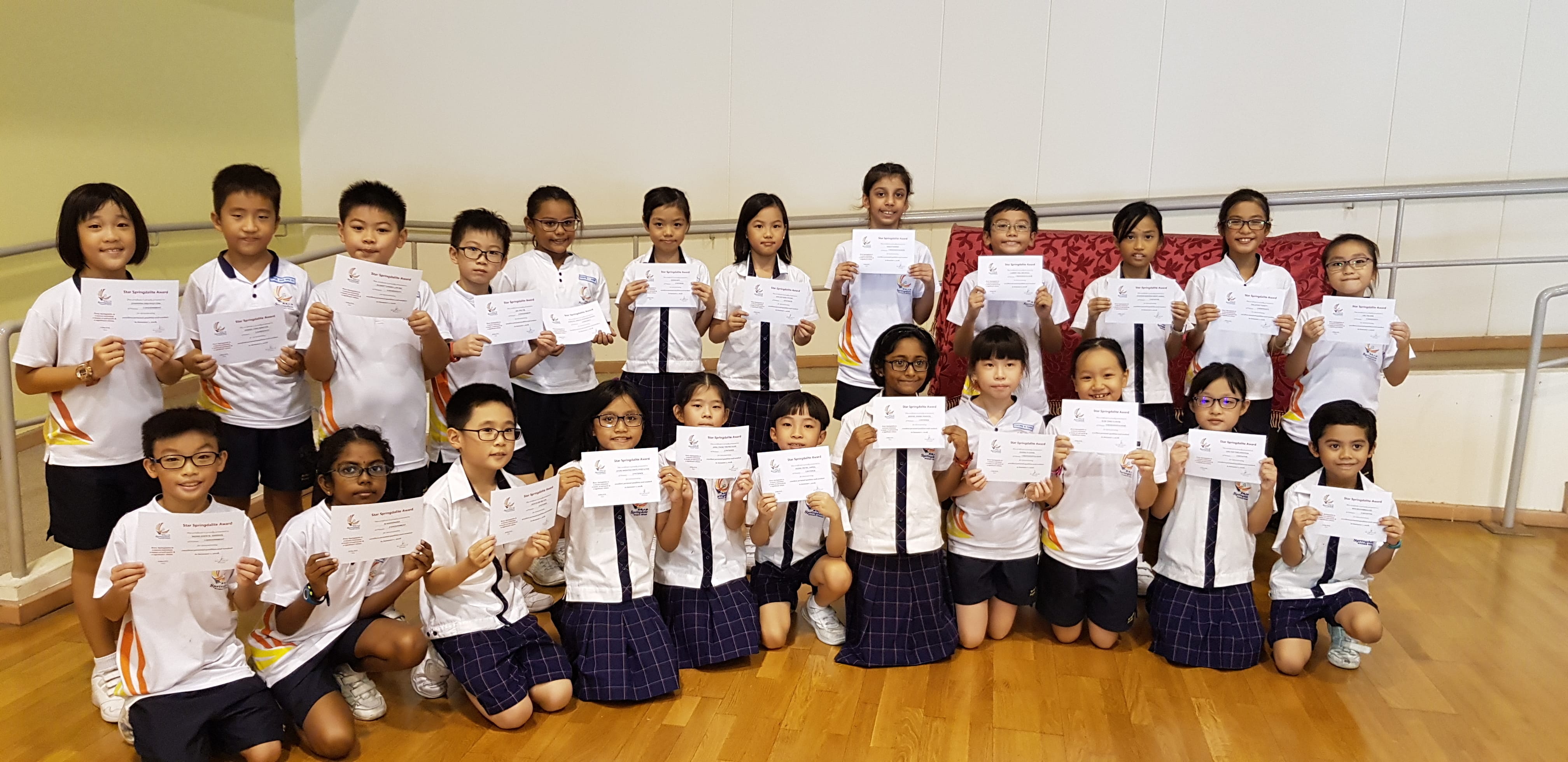 Group of young school children in white uniforms posing indoors, each holding a certificate.